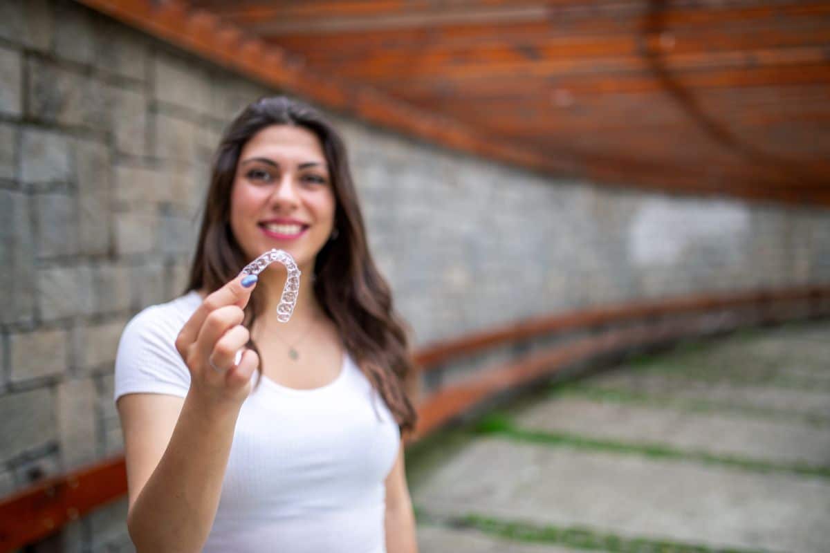 A woman smiling and holding a clear dental aligner outdoors.